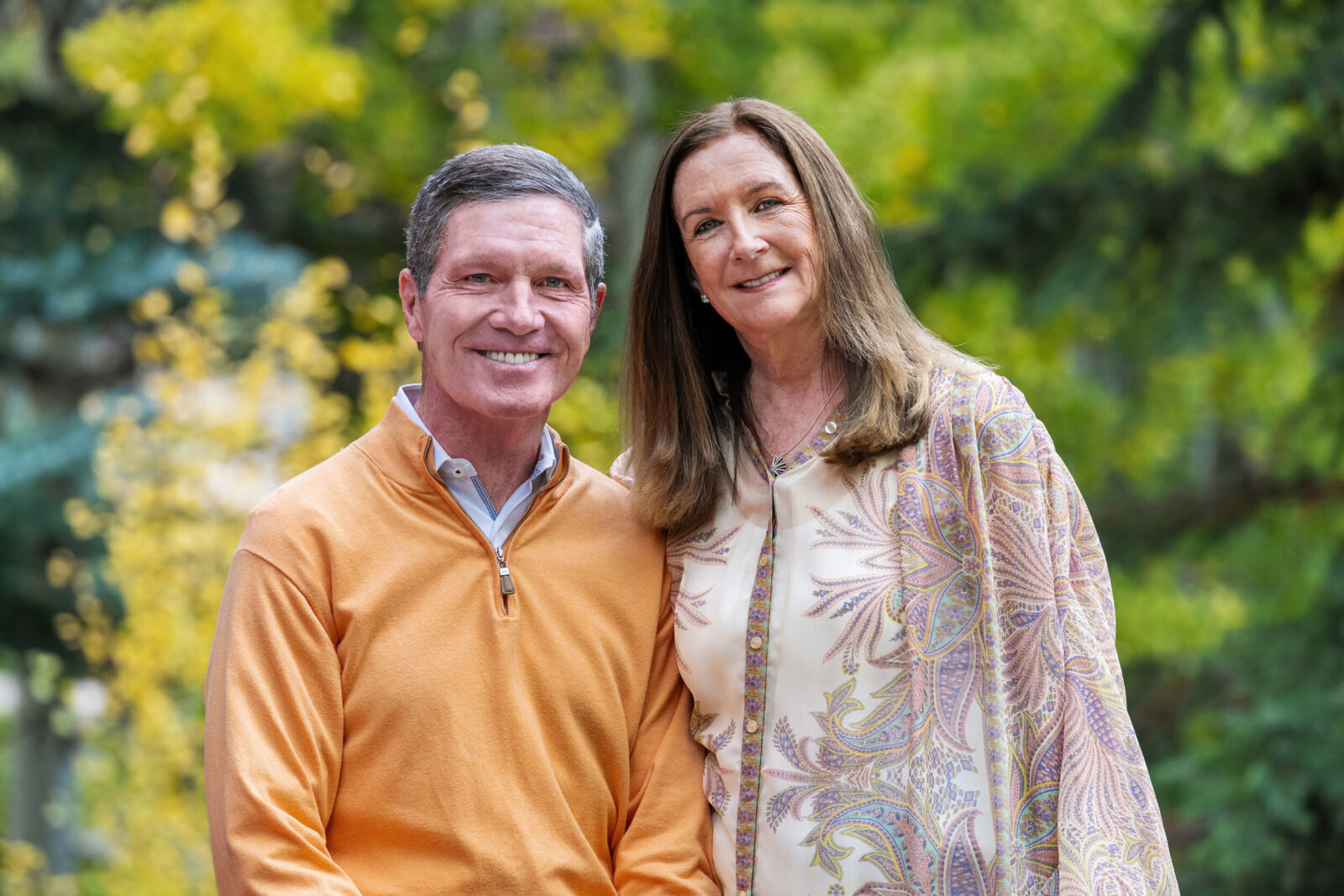 A man and woman pose for the camera and smile together with fall colors in the background