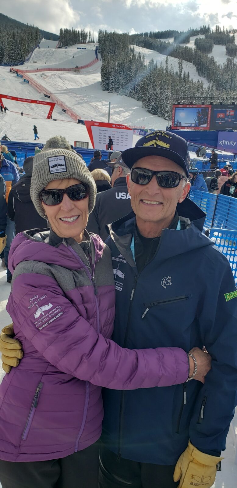 Debby and Harry Jasper smile together while wearing coats, beanies and sunglasses among a crowd of spectators at the Birds of Prey World Cup ski races with snow in the background